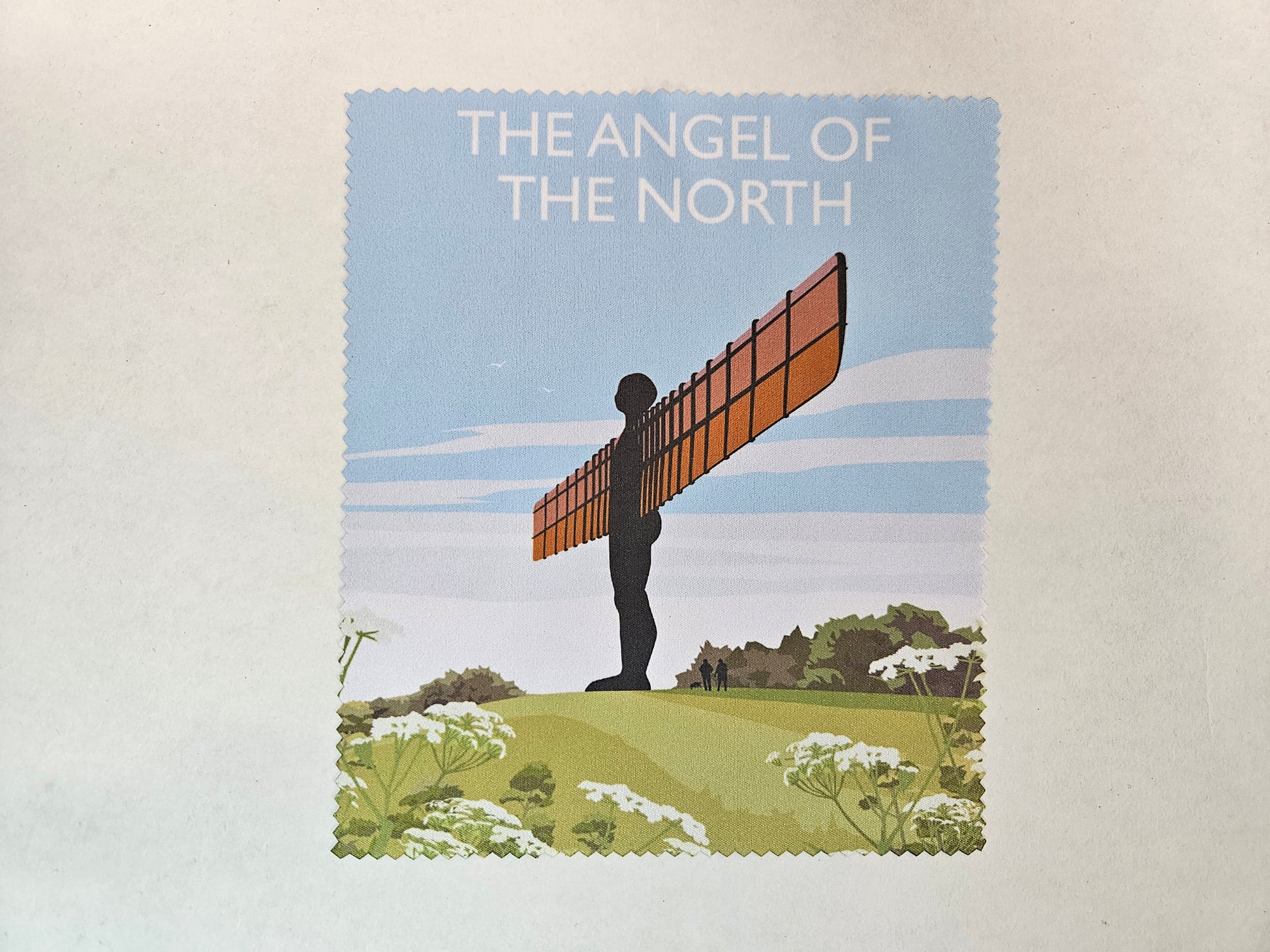A printed piece of cloth with zigzag edges showing a figure with outstretched arms or wings and the words the Angel of the North in white text. 