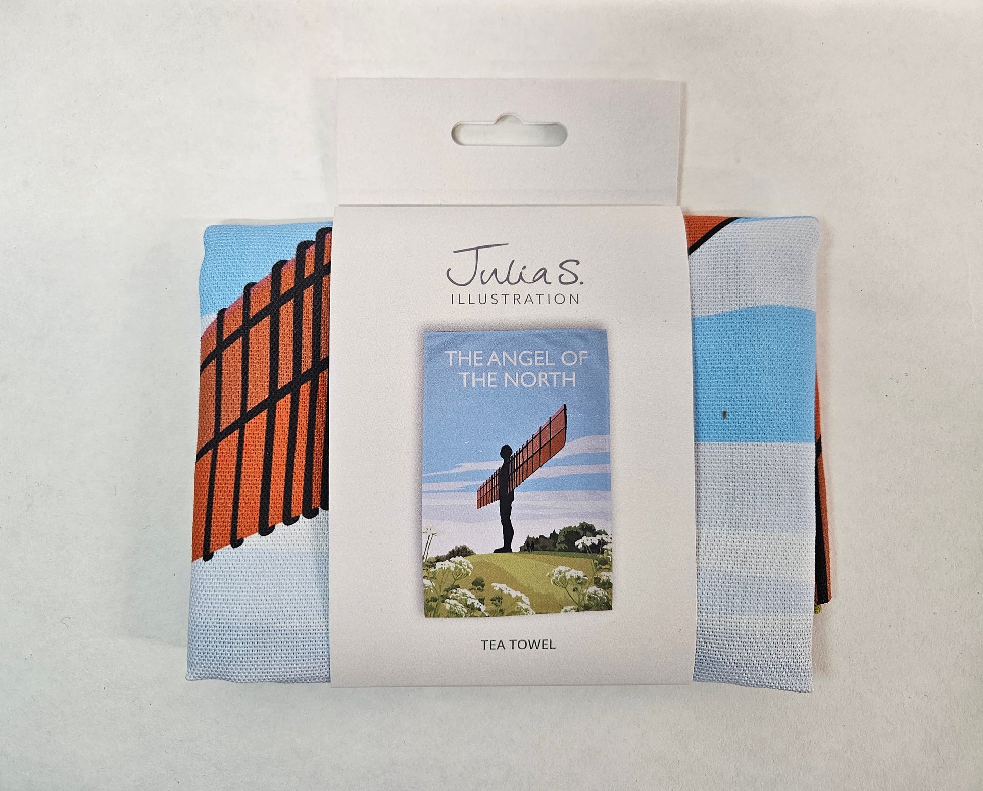 Julia S. Illustration tea towel in white packaging featuring an image of an dark orange figure with outstretched arm wings on a green hill with white flowers.