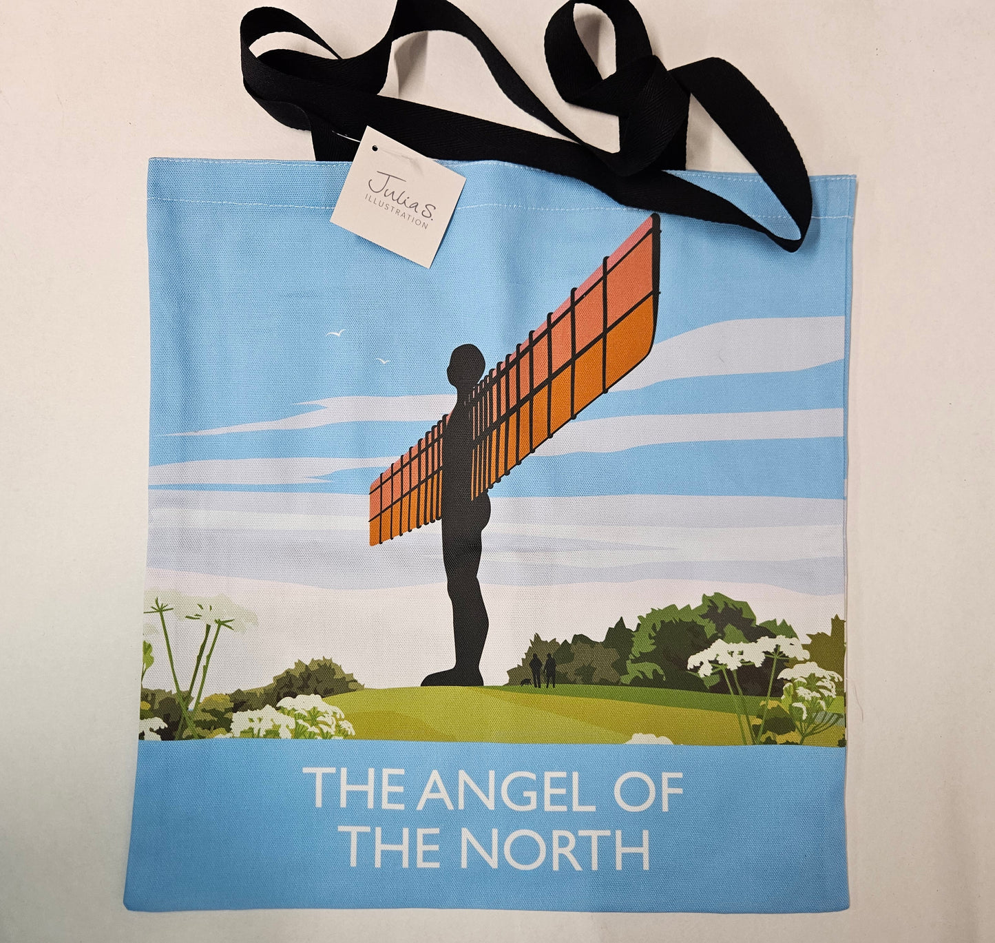A canvas tote bag featuring a graphic print of the Angel of the North sculpture on a blue sky background with green grass at the bottom and THE ANGEL OF THE NORTH below. The bag has long black shoulder straps. 