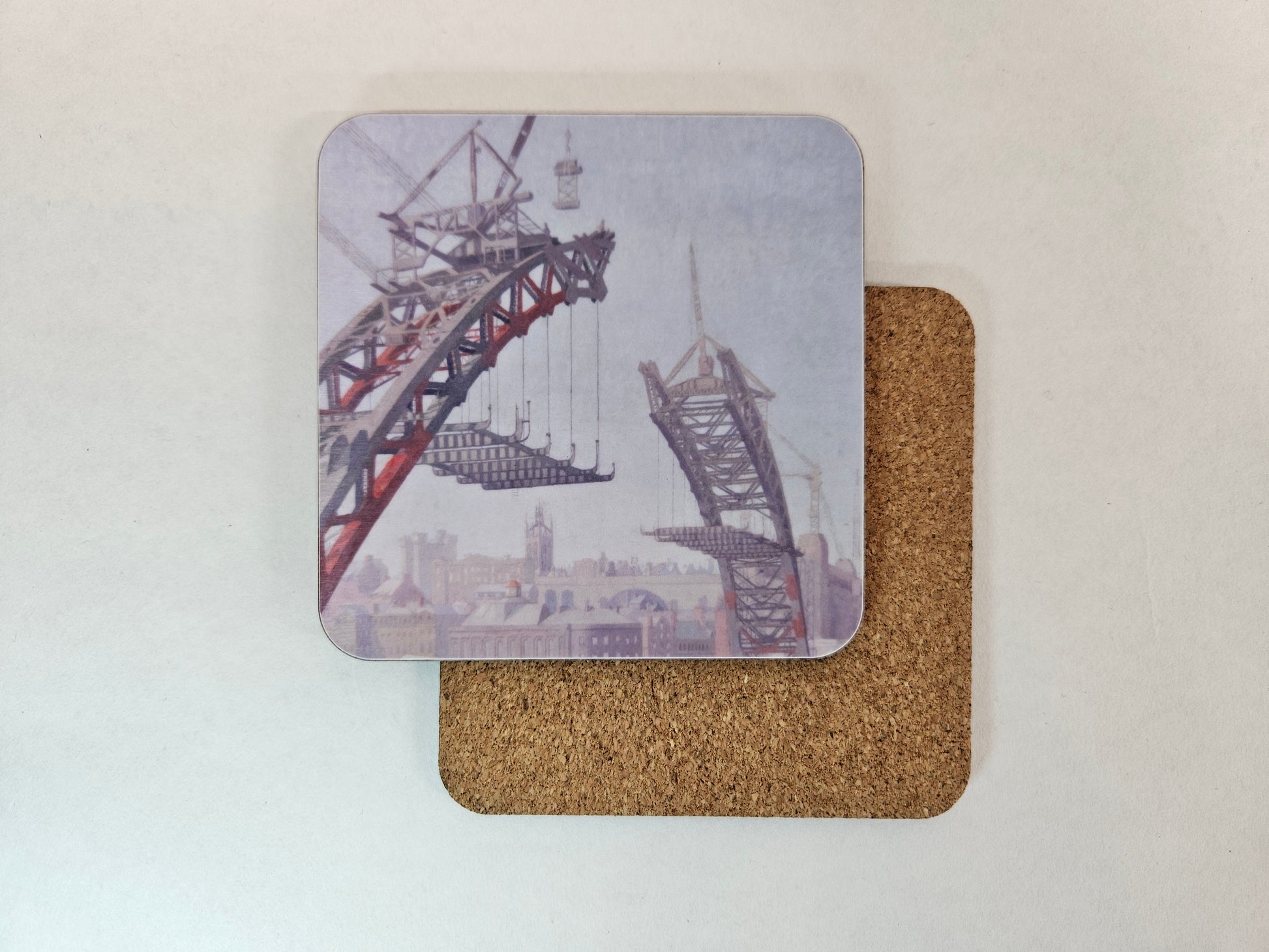 Two square coasters on a light surface, with one coaster showing a printed industrial crane and cityscape design and the other showing the natural brown cork backing.