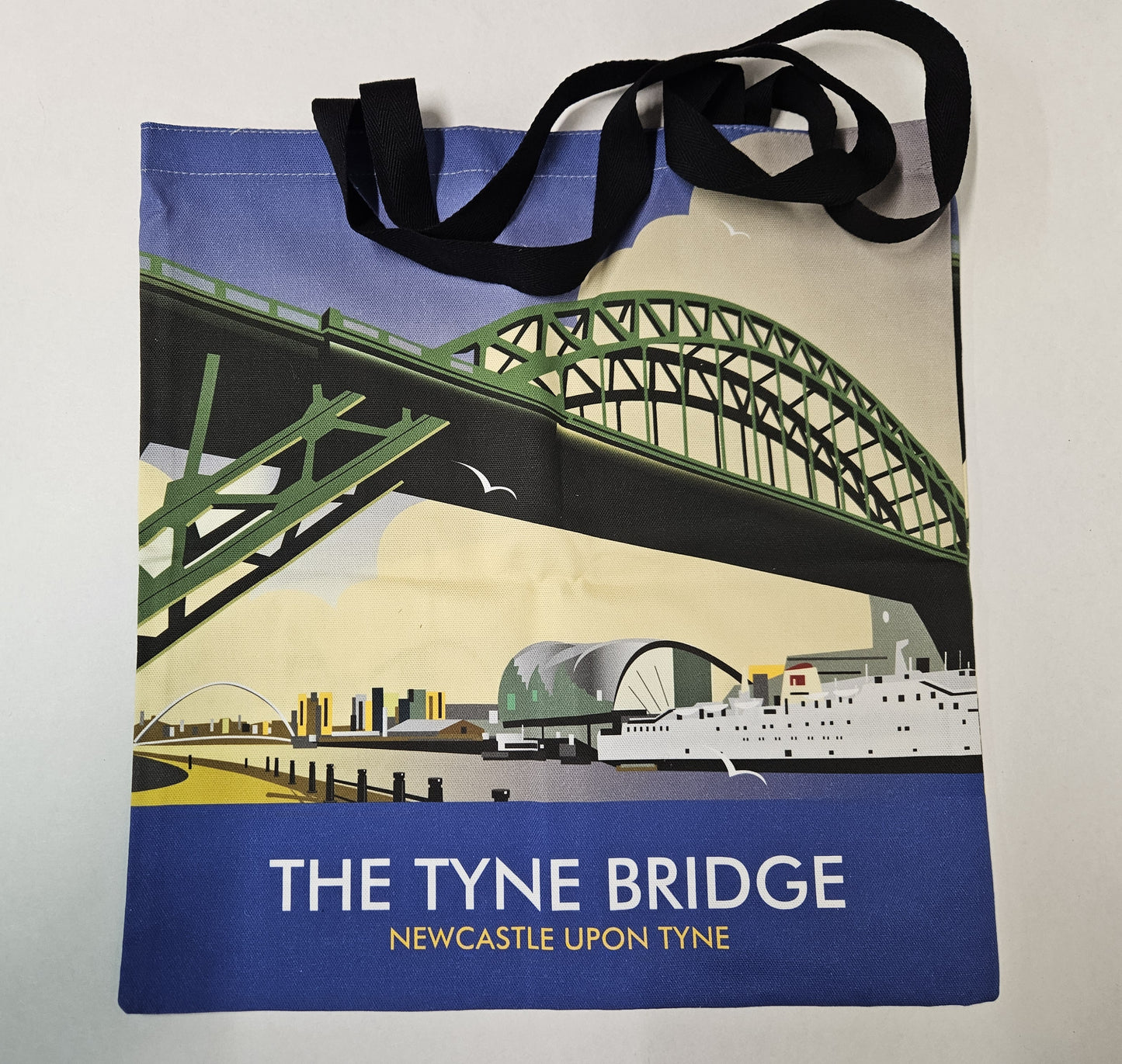 A tote bag with black long handles and a colourful image of the green bridge with THE TYNE BRIDGE, NEWCASTLE UPON TYNE with White boat, bridges and buildings in the background. 