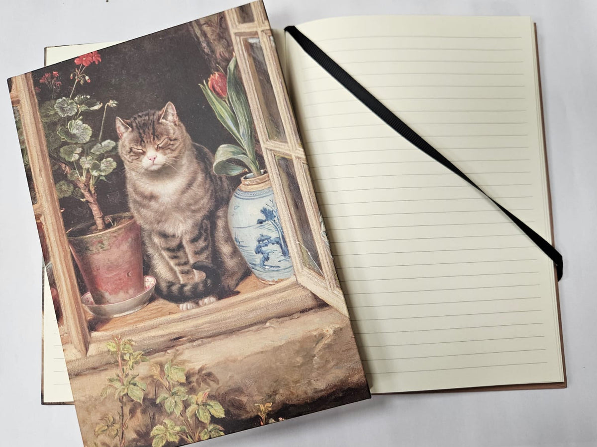 Notebook with a printed illustration of a cat sitting by a window next to potted plants, with open journal behind with cream paper and black ribbon page marker. 
