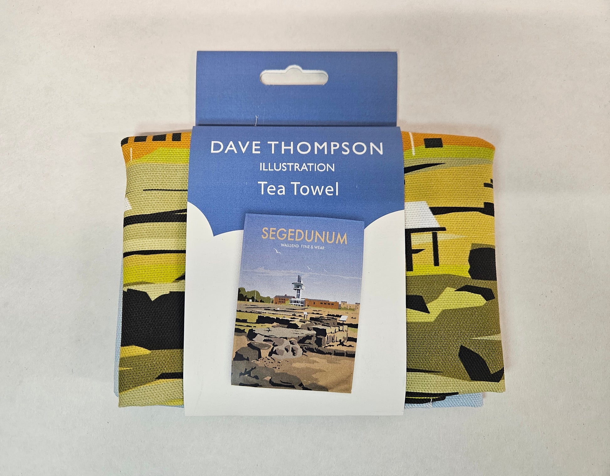 A folded tea towel packaged with a blue cardboard header card that reads 'DAVE THOMPSON ILLUSTRATION Tea Towel' and shows a small 'SEGEDUNUM' image that is depicted on the tea towel. 