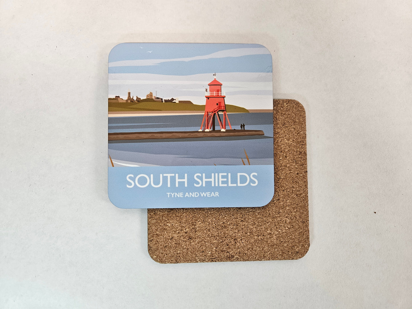 A square cork coaster with rounded corners lies on a light beige surface, partially covering another coaster. The top coaster features a printed illustration of a red lighthouse on a pier over water, with the text 'SOUTH SHIELDS' and 'TYNE AND WEAR' on a pale blue background.