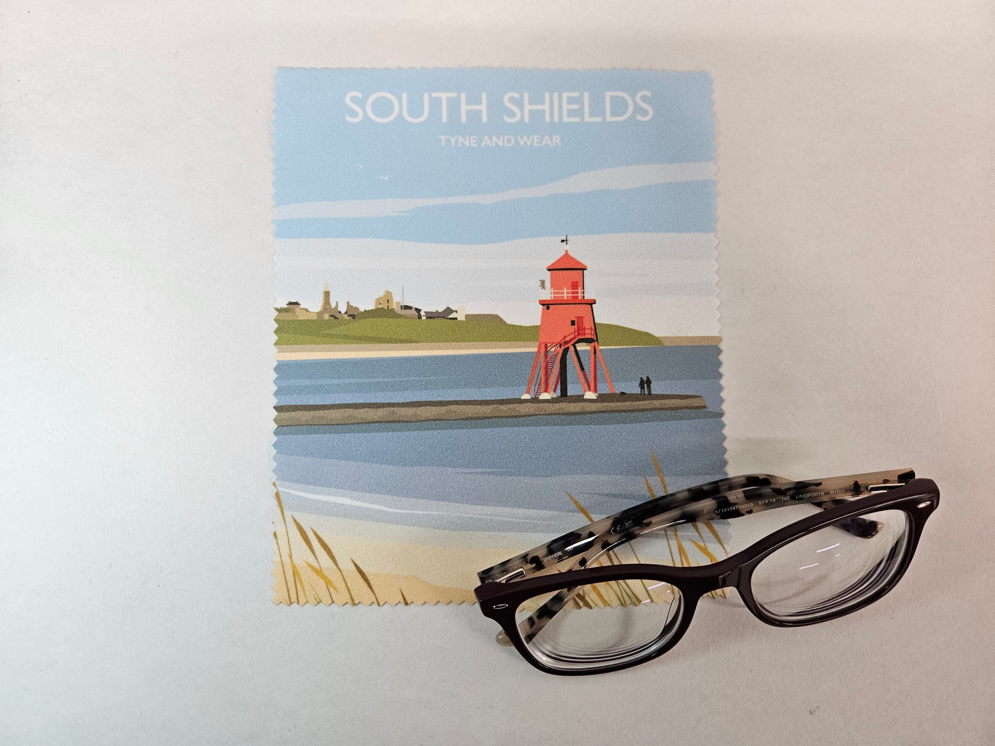 A pair of black cat-eye eyeglasses with patterned temples rests on top of rectangular printed fabric featuring a coastal lighthouse illustration and the text 'SOUTH SHIELDS TYNE AND WEAR'.