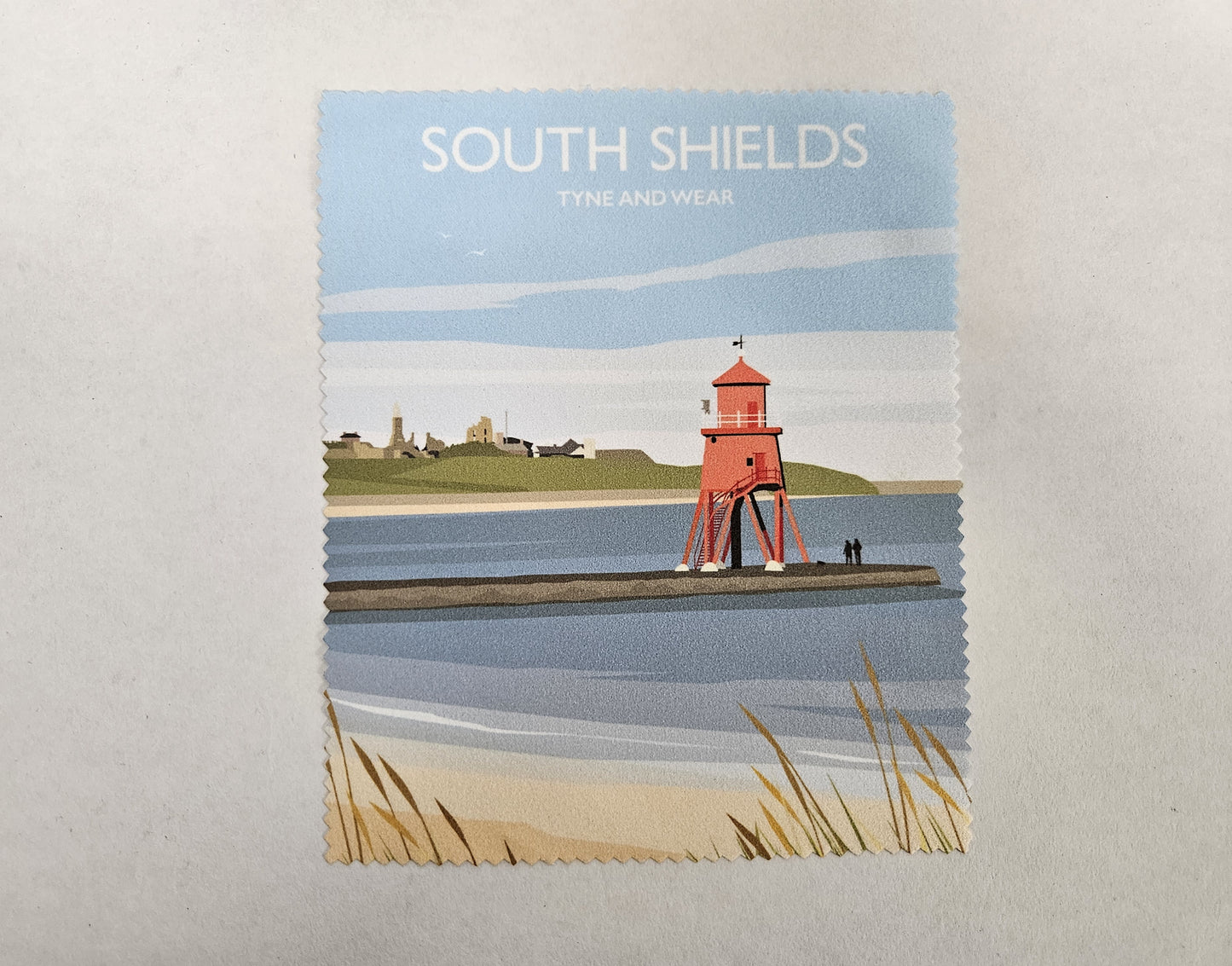 A rectangular art print with zig-zag edges showing a coastal scene labelled 'SOUTH SHIELDS TYNE AND WEAR.' The illustration features a red lighthouse on a pier over blue water, with pale sand, grasses, and buildings in the distance under a light blue sky.