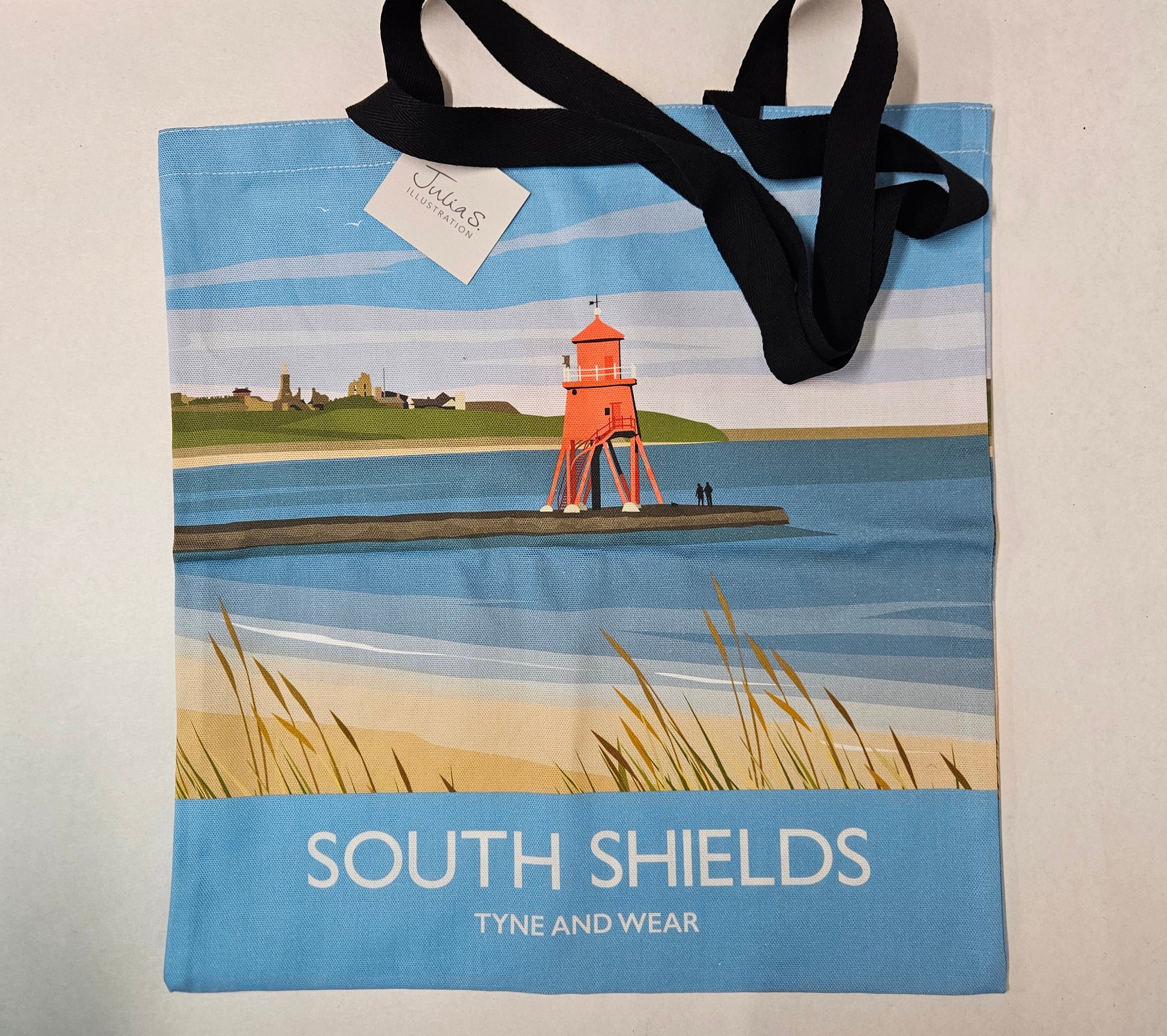 A canvas tote bag with black shoulder straps, featuring a printed illustration of a red lighthouse on a pier over blue water, with the text 'SOUTH SHIELDS TYNE AND WEAR' on a light blue band at the bottom. A small white paper tag reading 'Tyne and Wear' is attached near the top.